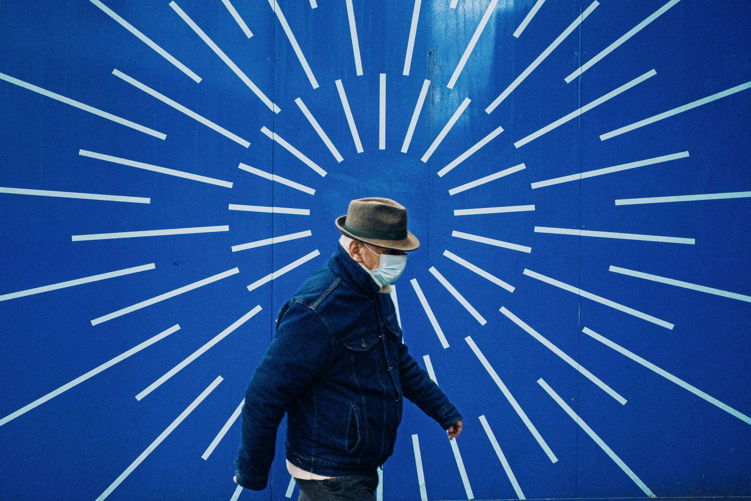Man wearing a face mask and hat walks past a bold blue wall with white sunburst pattern.