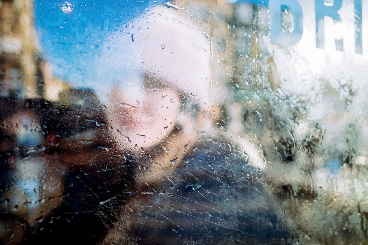 Person seen through a rain-covered window with water droplets, creating a blurred, moody urban scene.
