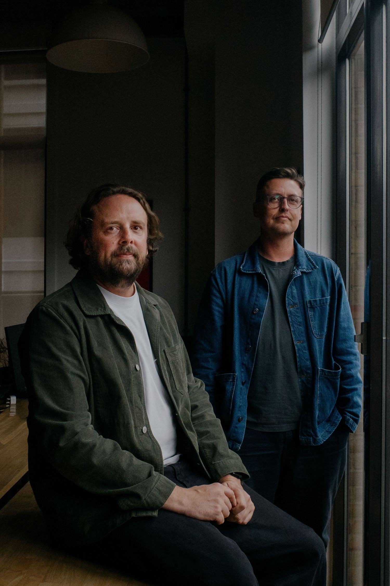 Two bearded men in casual jackets pose near a window in a dimly lit modern office space.