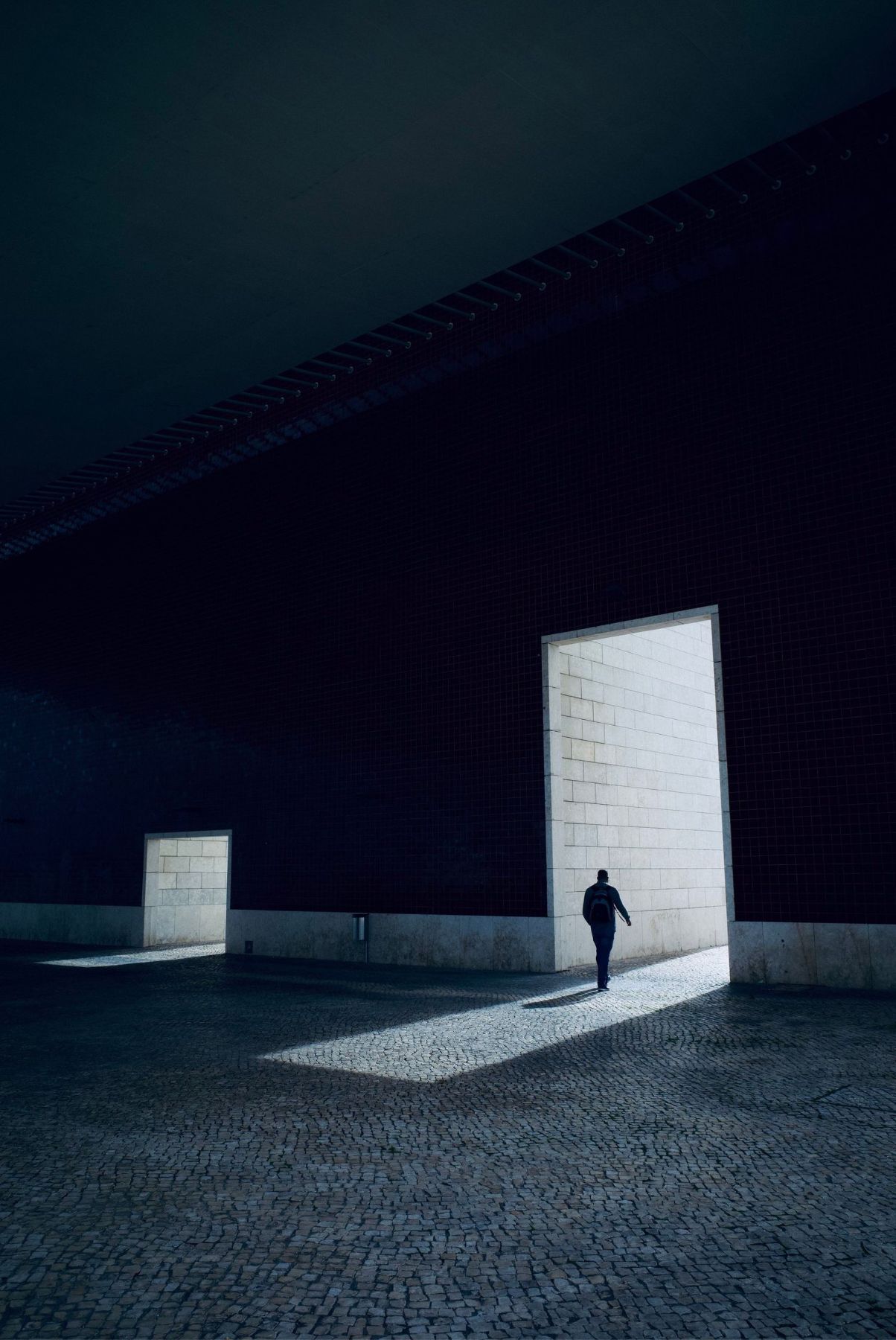 A lone figure walks toward a bright doorway in a dark cobblestone courtyard at night.