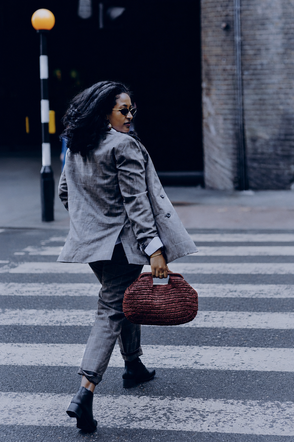 Woman in gray blazer and trousers crossing a zebra crossing while carrying a red woven handbag.