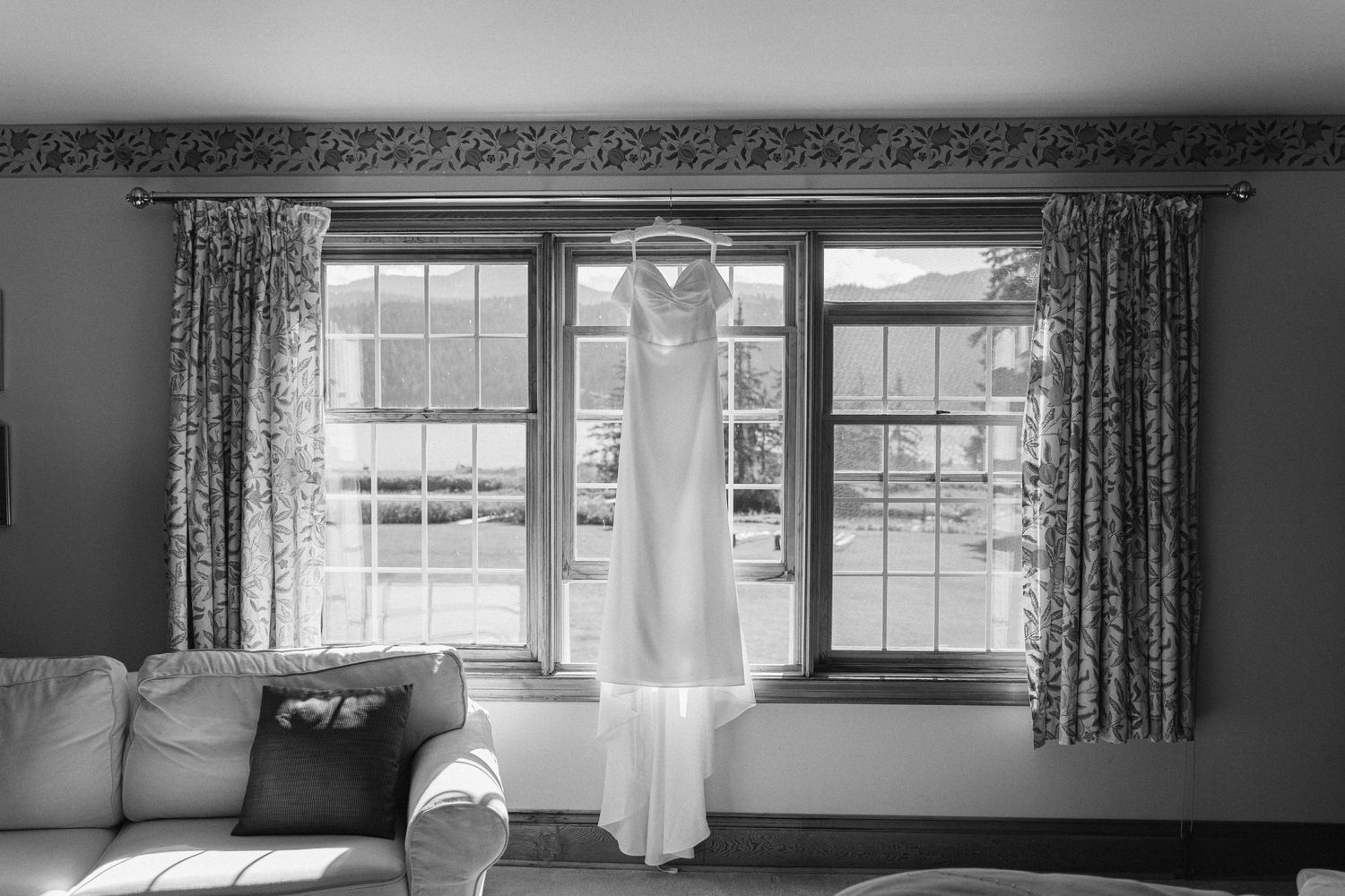 A white wedding dress hangs elegantly in front of three sunlit windows with floral curtains.