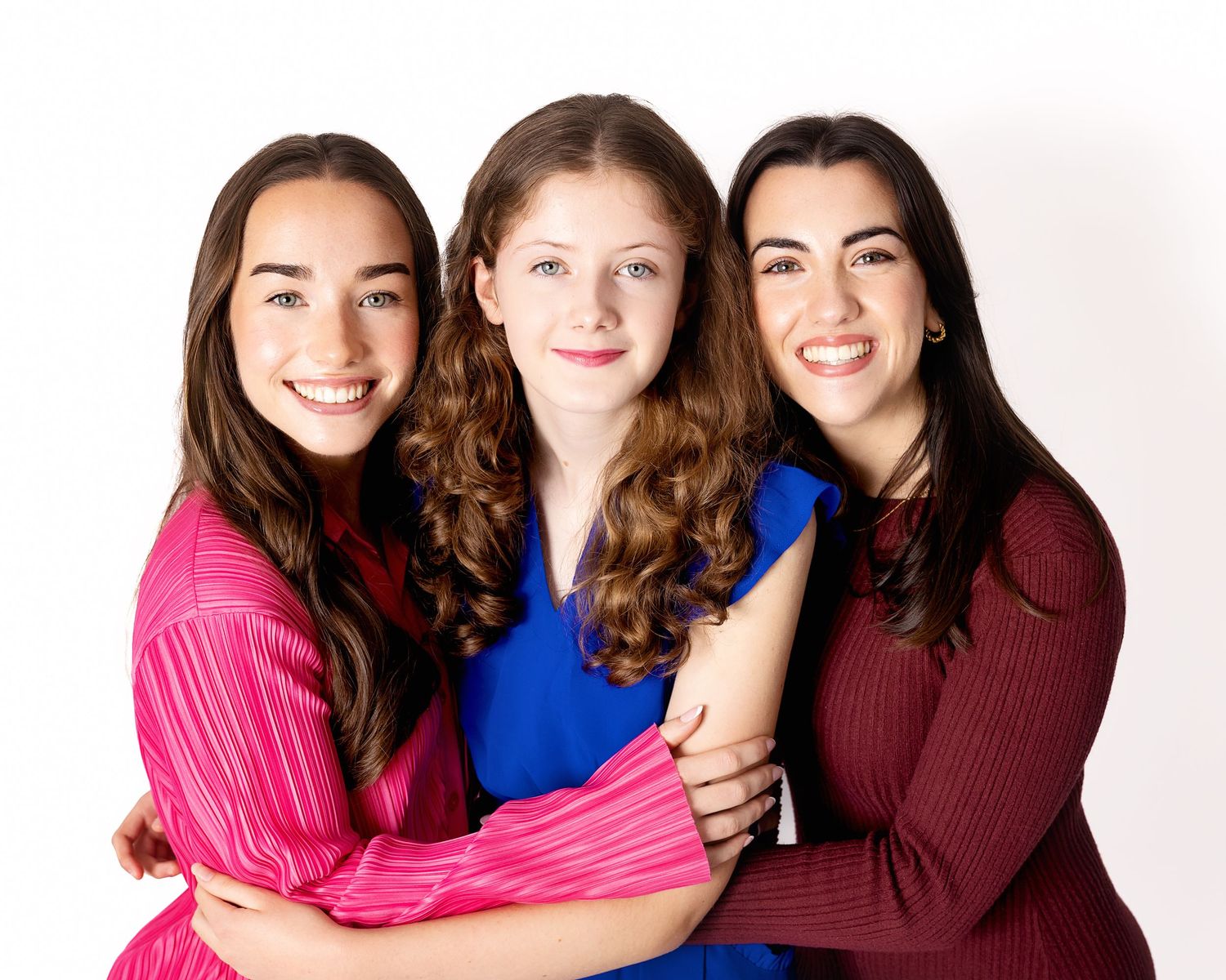 Three smiling friends wearing bright pink, blue and burgundy sweaters pose together against a light background.