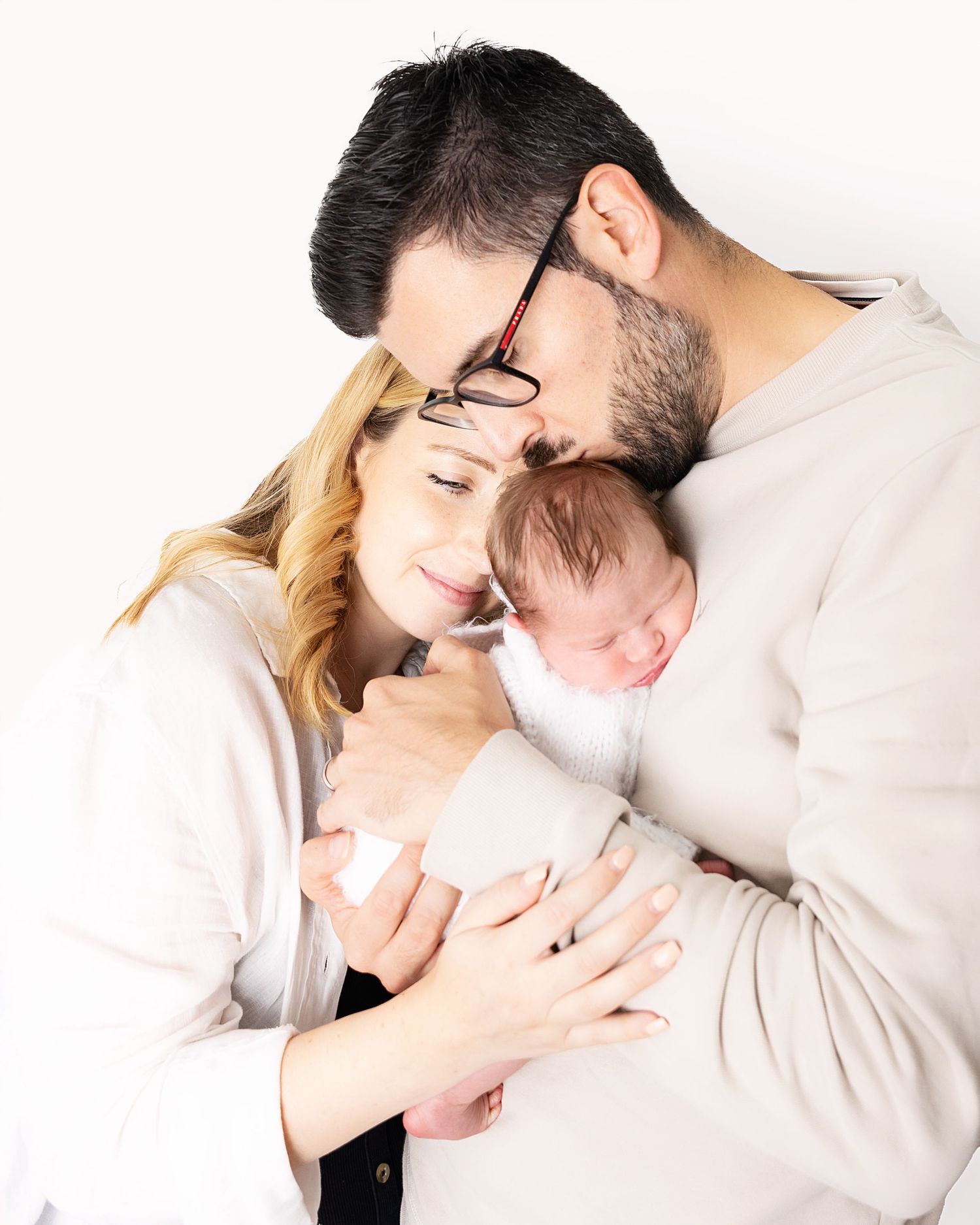 A tender family moment captured in white as parents lovingly embrace their sleeping newborn baby.