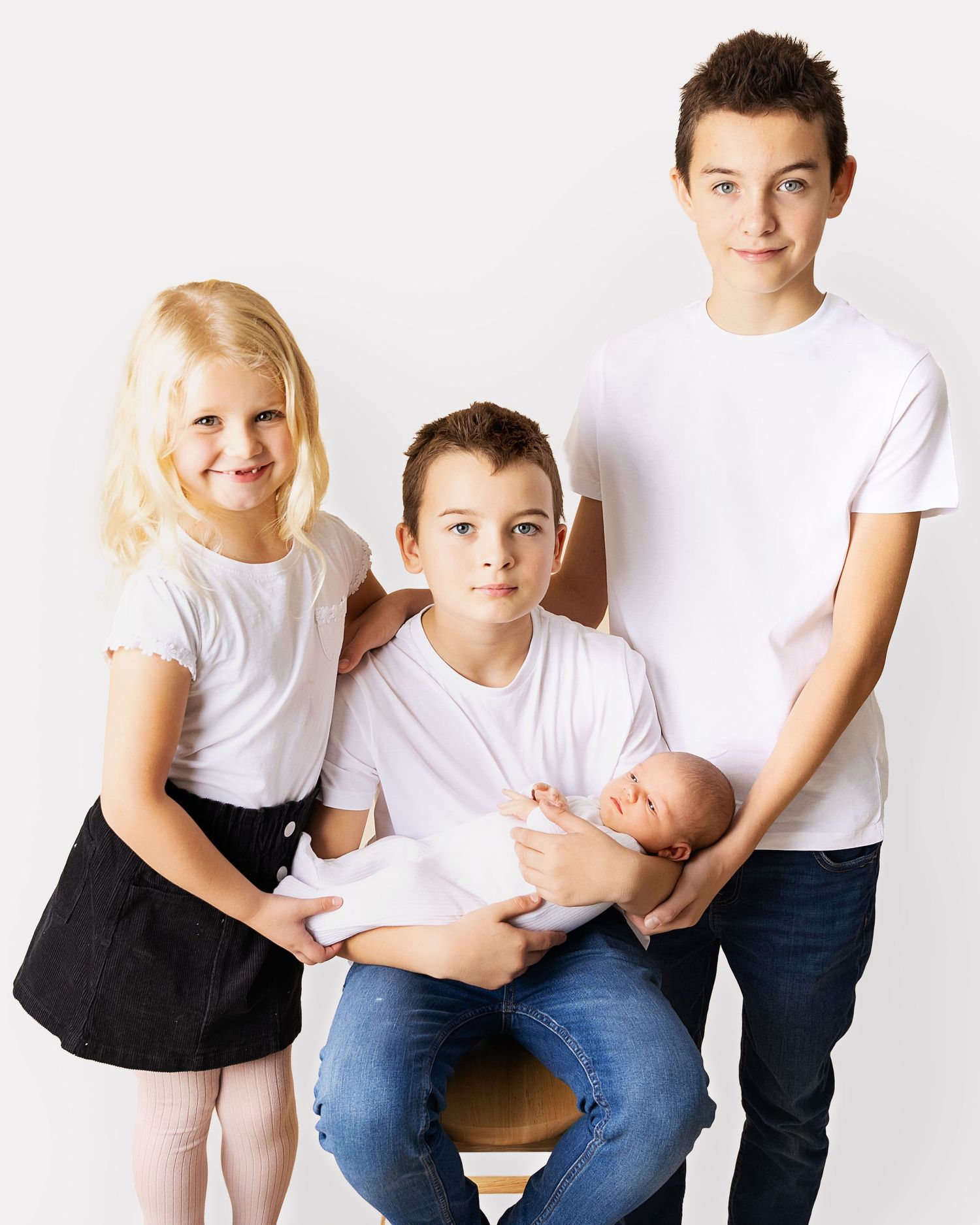 Three siblings in matching white t-shirts pose together while holding their newborn baby sibling against white background.