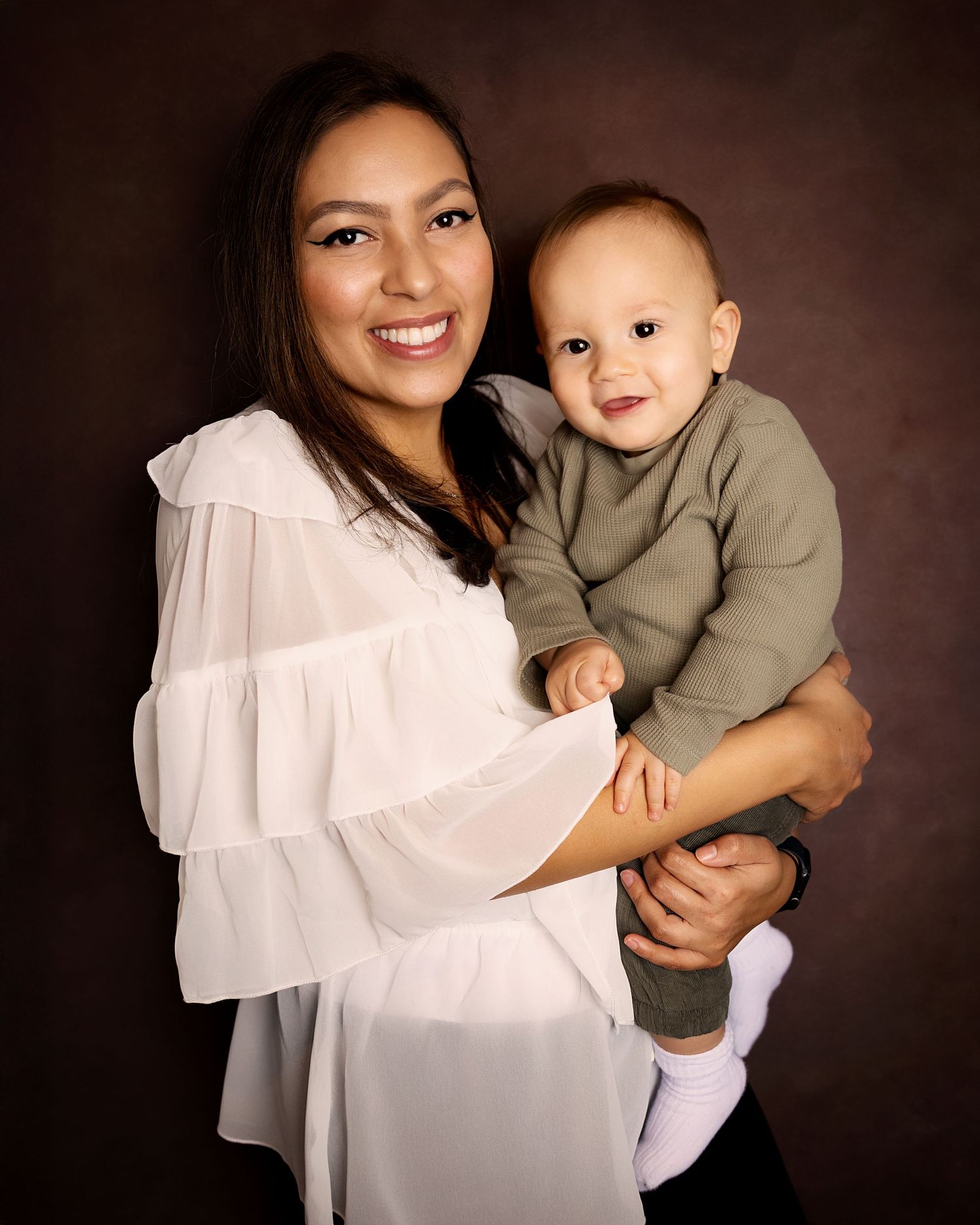 A feminine figure in a white ruffled blouse holds a smiling baby wearing a gray outfit against a dark brown backdrop.