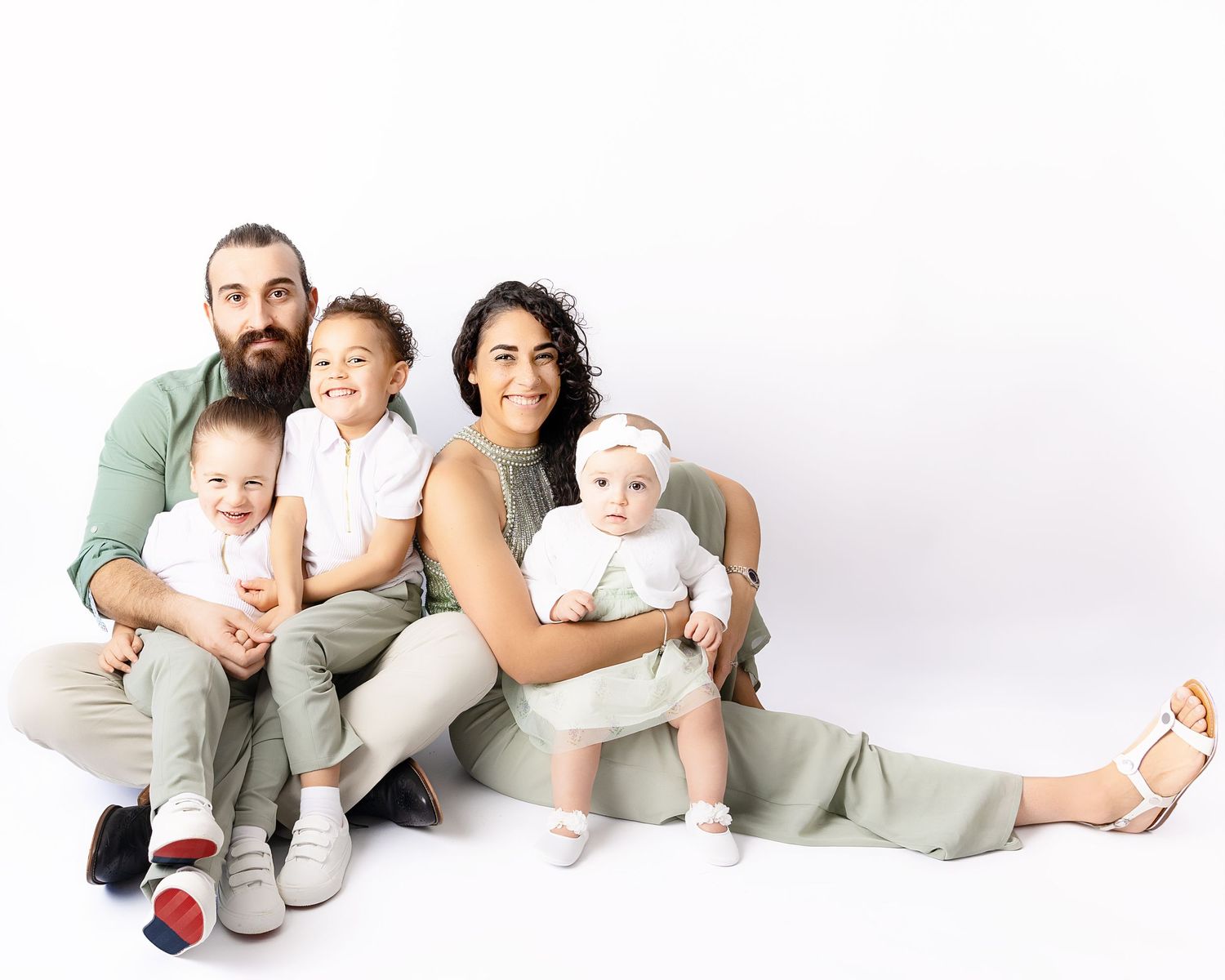A family wearing coordinated neutral outfits sitting together on a white background for a portrait session.