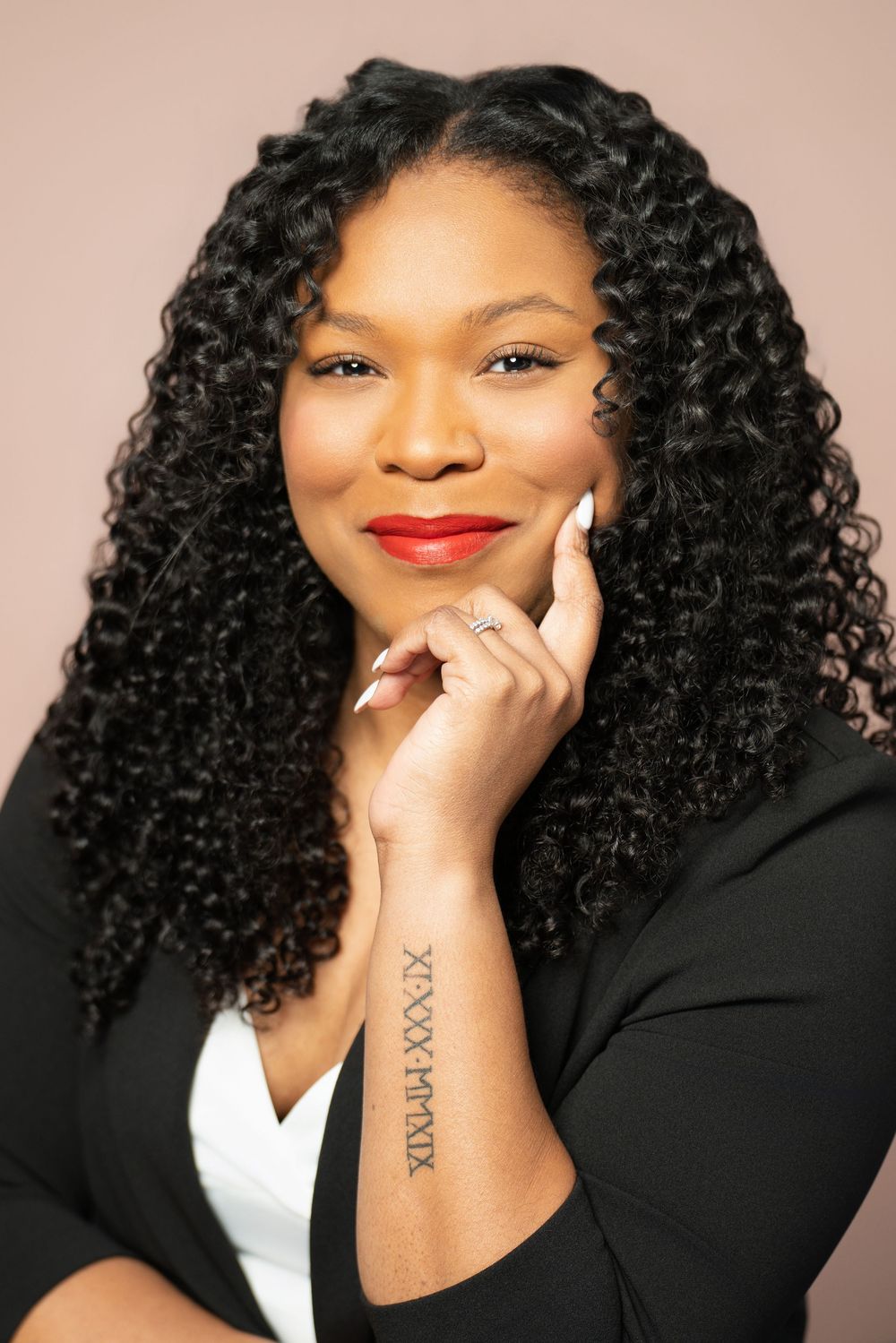 Professional portrait of a person with curly black hair, red lipstick and a black blazer against a neutral background.
