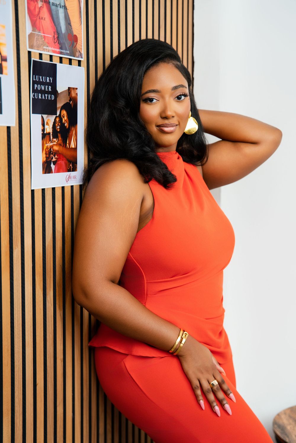 Woman in vibrant coral sleeveless dress poses confidently against wooden slat wall with framed artwork.