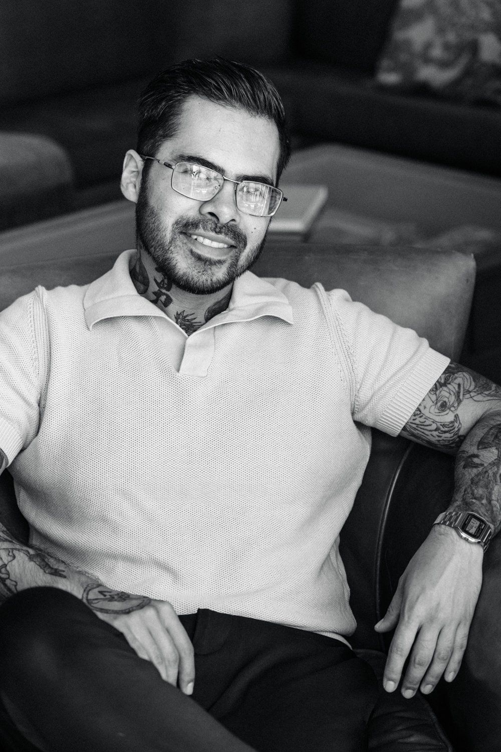 A casual black and white portrait of a person with tattoos wearing glasses and a polo shirt.