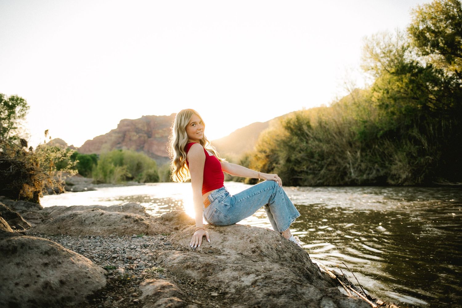 Woman in black top and white wide-leg pants sitting on rocks at sunset with mountain backdrop.