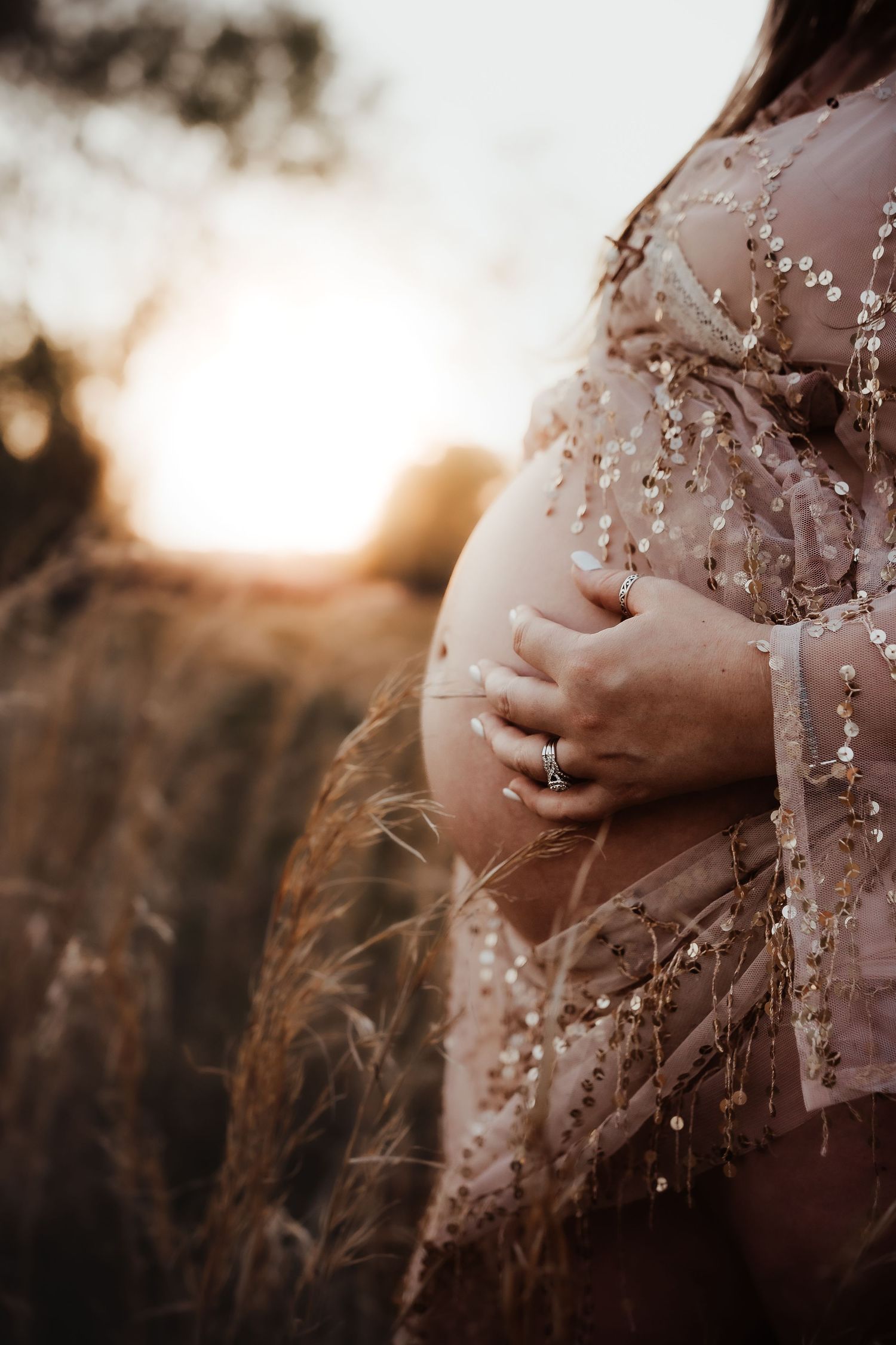 Pregnant belly draped in a sequined dress with golden light behind in a field of golden grass