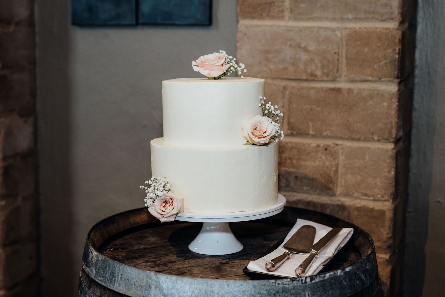Elegant white wedding cake decorated with delicate blue and white flowers sits on a wooden cake stand with twinkle lights in background.