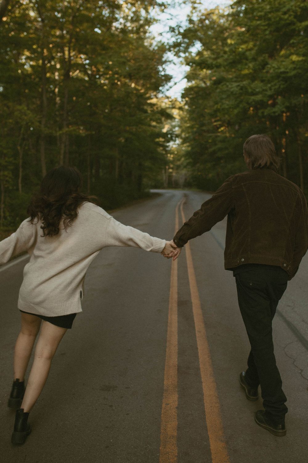A couple holding hands while walking down a tree-lined road surrounded by lush green forest.