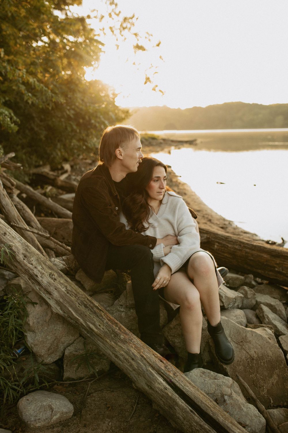 A couple sitting on rocks by a lakeside at golden hour, embracing in a serene natural setting.