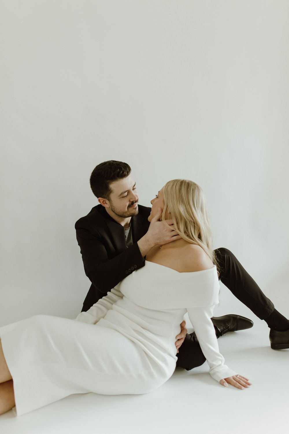Couple in elegant attire sitting on the floor gazing at each other in a bright white studio.