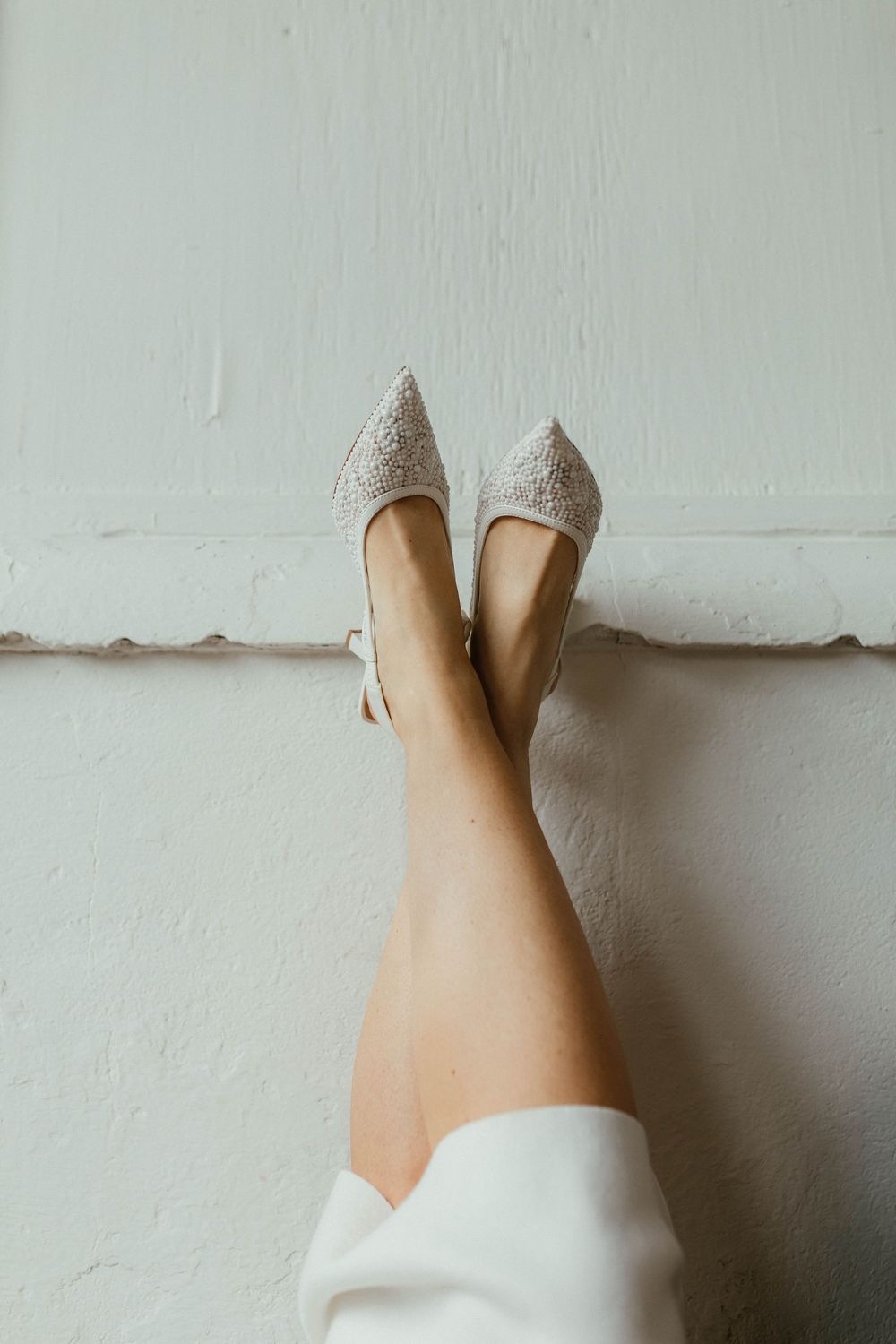 Close-up of a woman's legs raised against a white wall wearing sparkly pointed-toe heels.