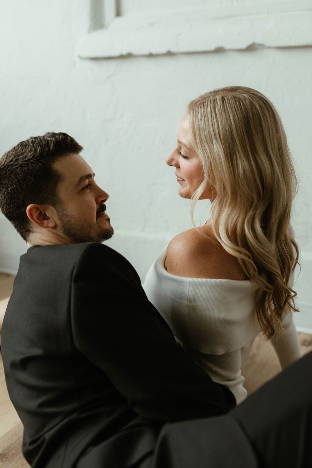 Couple sitting on the floor sharing an intimate glance, woman in off-shoulder white top.