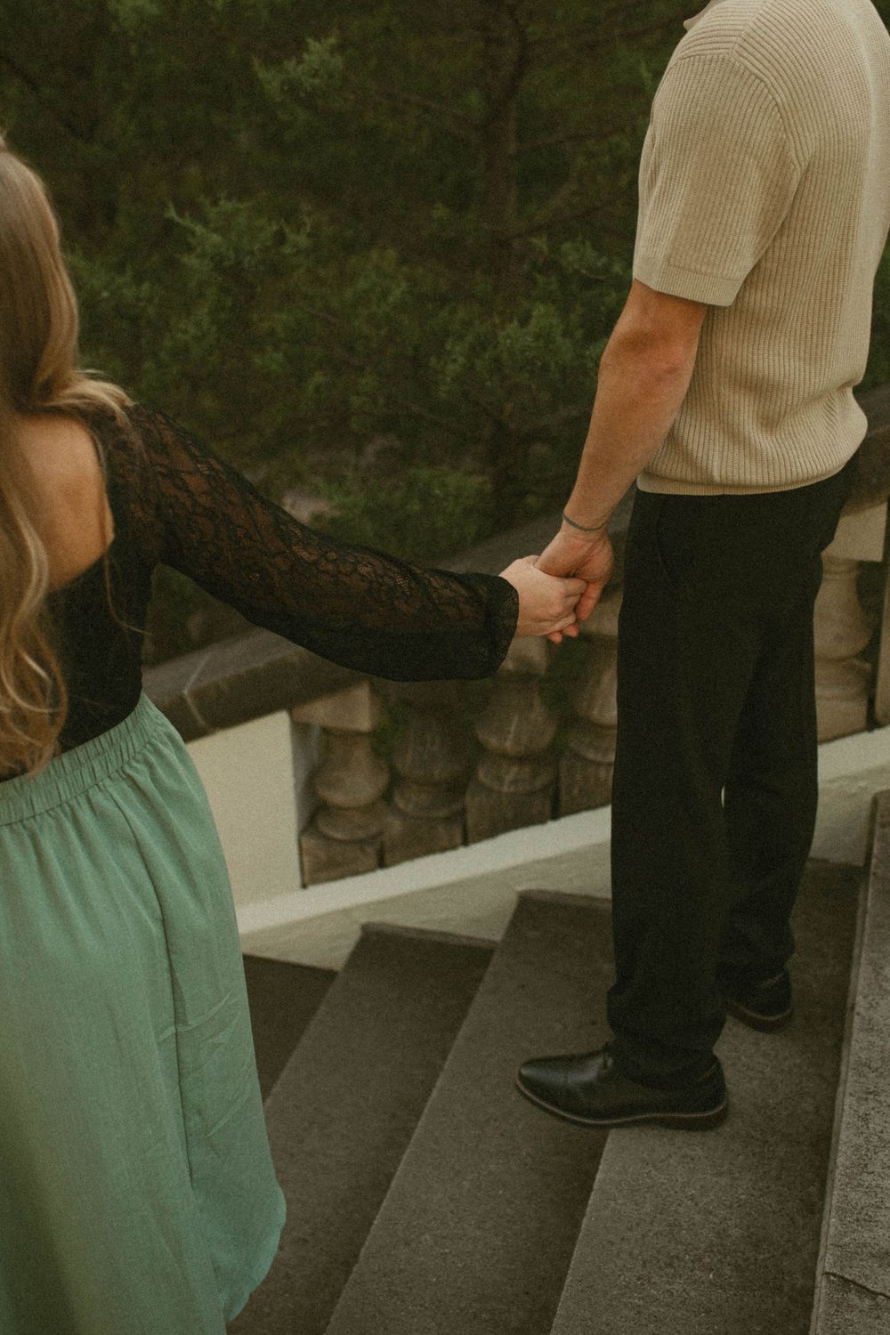 Couple holding hands on stone steps, woman in black lace top and teal skirt.
