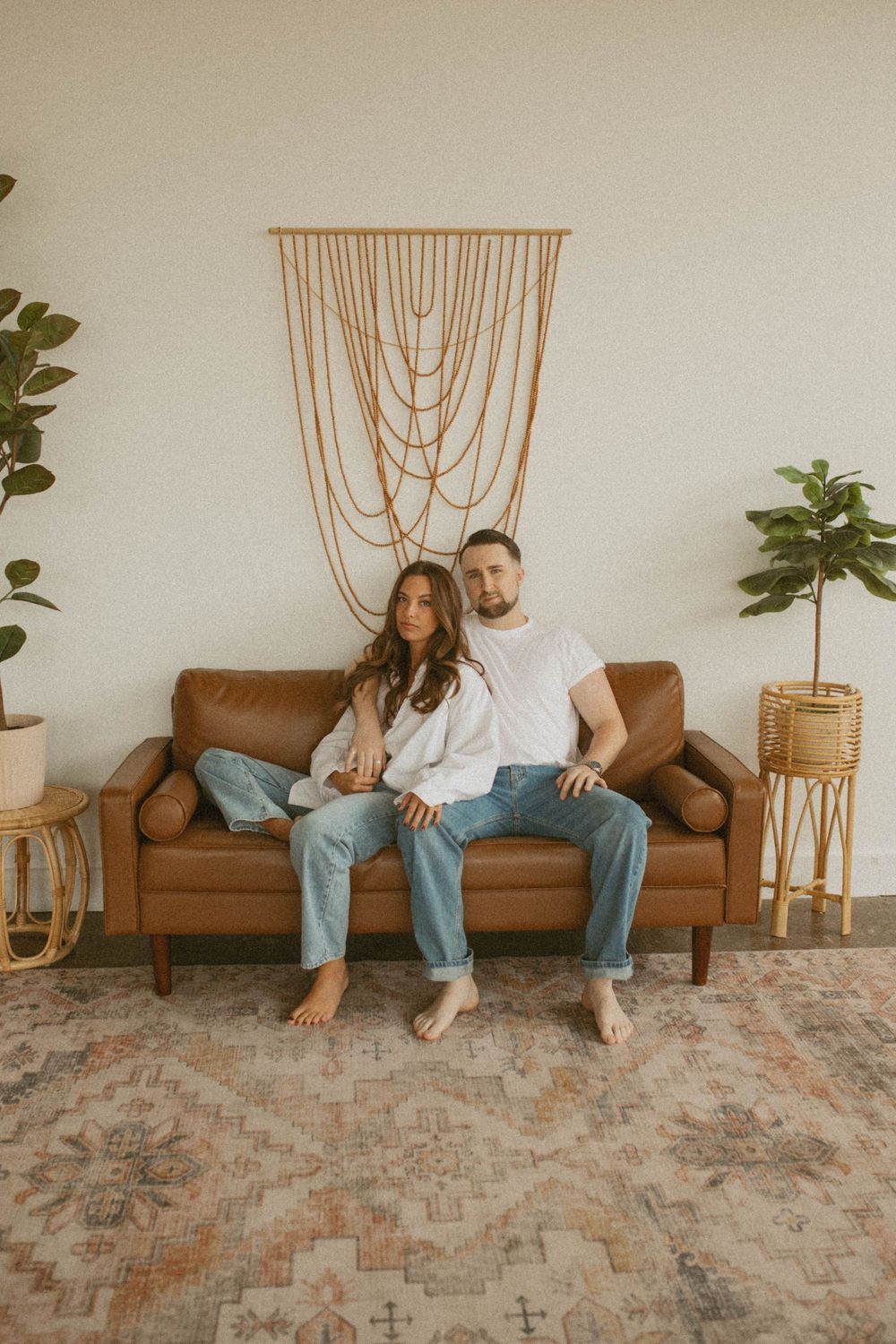 A couple sitting together on a brown leather sofa in a boho-styled living room with macrame wall art and plants.