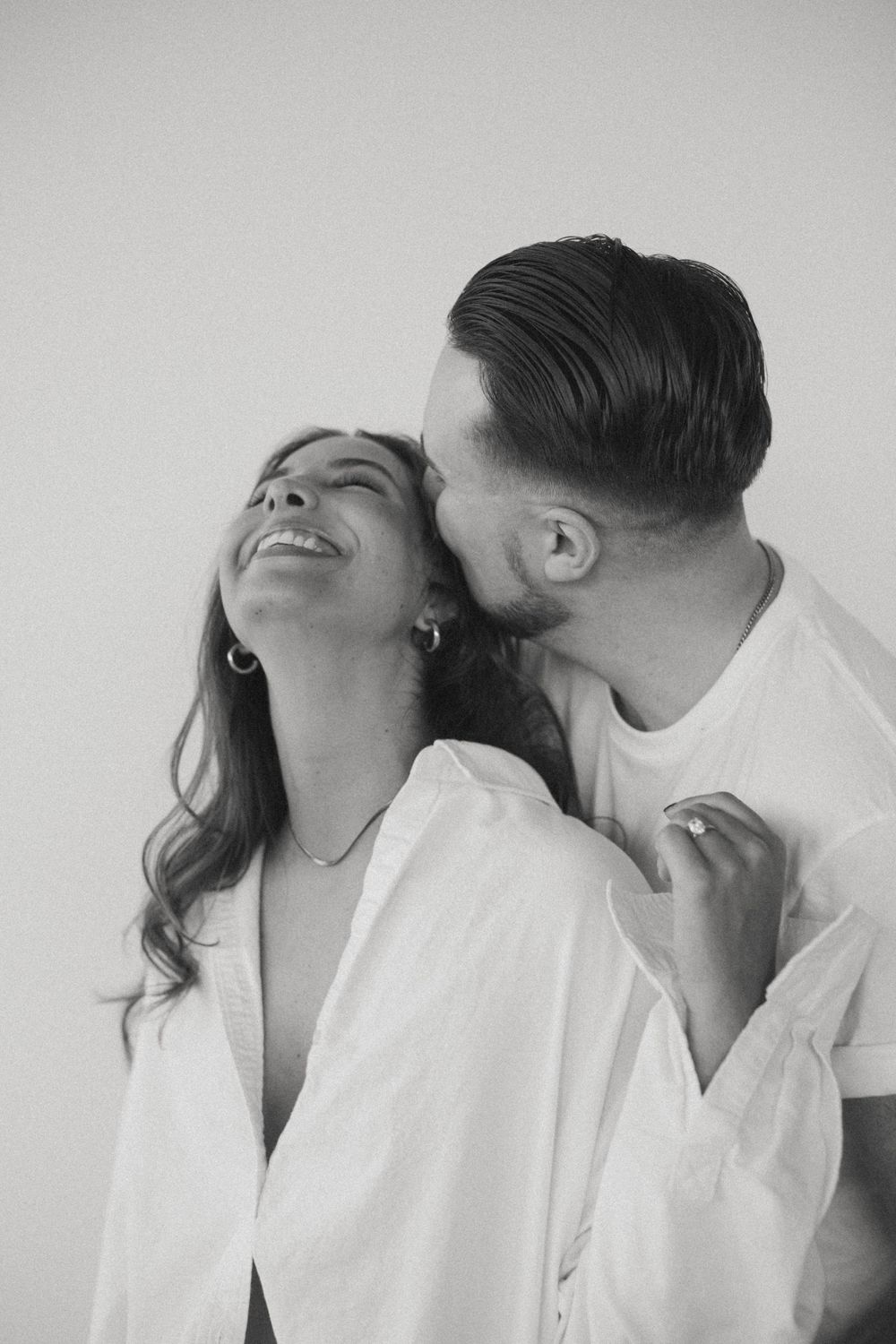 Black and white photo of a man kissing a smiling woman on the neck in matching white outfits.