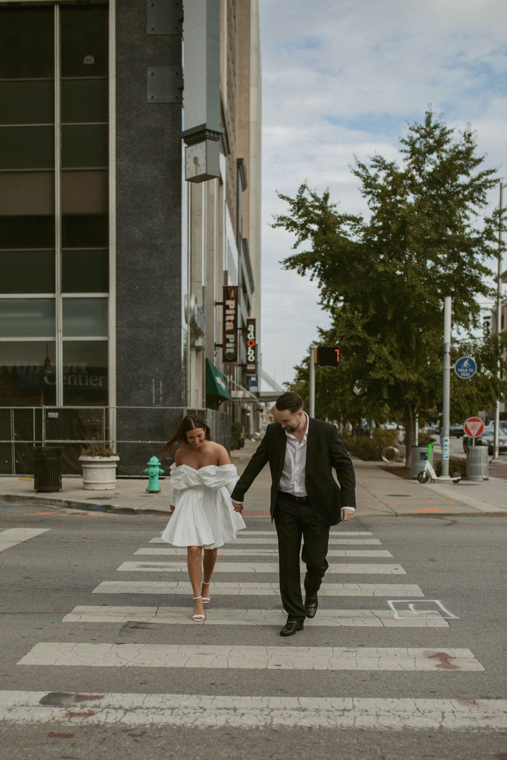 Couple in wedding attire holding hands while crossing a city street crosswalk together.
