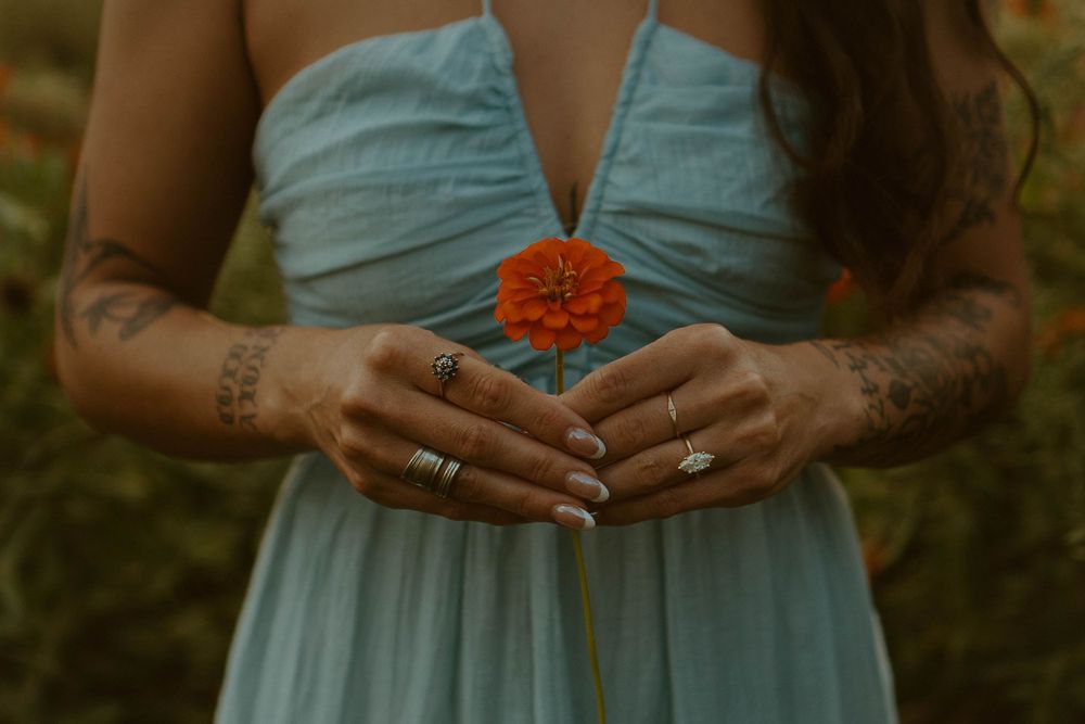 Woman in blue dress holding a red zinnia flower, showing tattooed arms and rings.