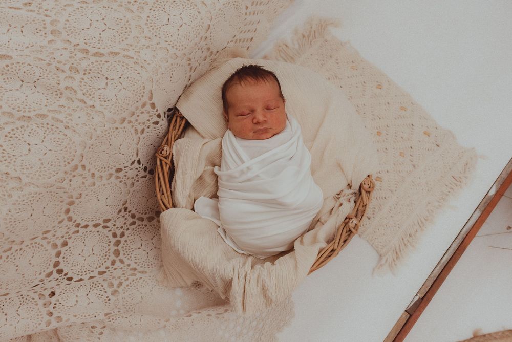Swaddled newborn sleeping peacefully on cream-colored textured blanket in woven basket.