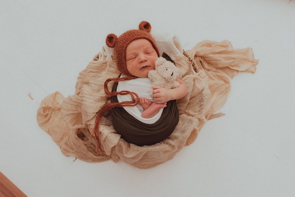Overhead view of sleeping newborn wearing brown bear hat laying on swirling beige fabric.