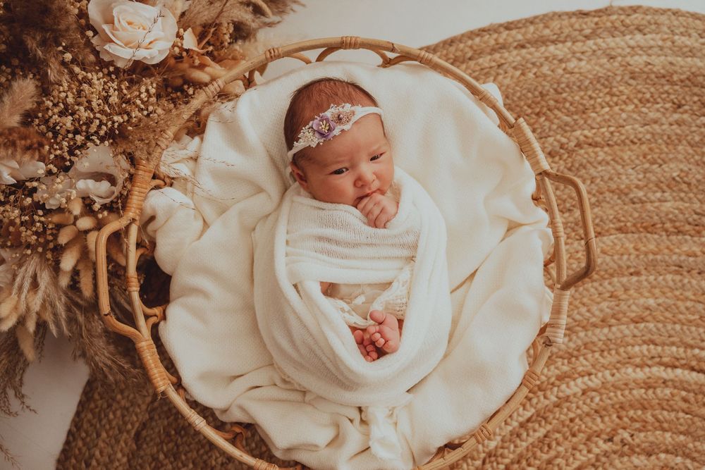 Newborn baby wrapped in white swaddle with lace headband sleeping in woven basket with dried floral decorations.