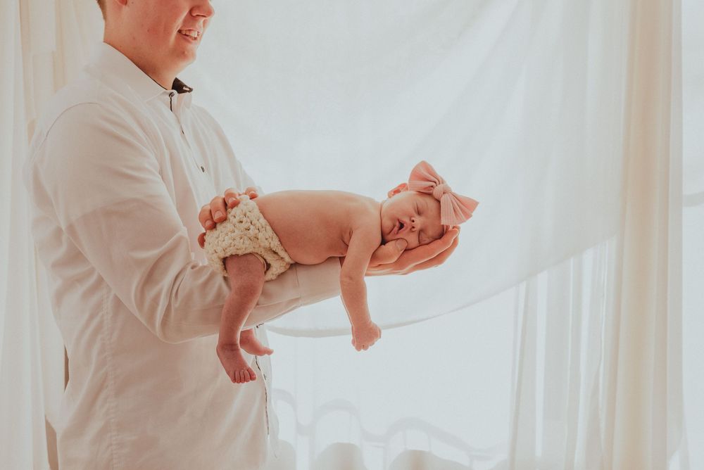 A person wearing a white shirt holds a sleeping newborn baby against a light background with soft natural lighting.