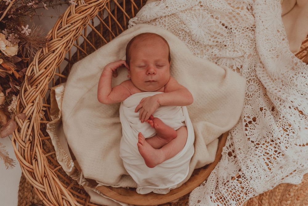 Sleeping newborn wrapped in white swaddle blanket rests peacefully in woven basket with lace details.