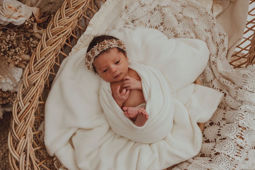 Newborn baby wrapped in soft white blanket sleeping peacefully in woven basket with decorative headband.