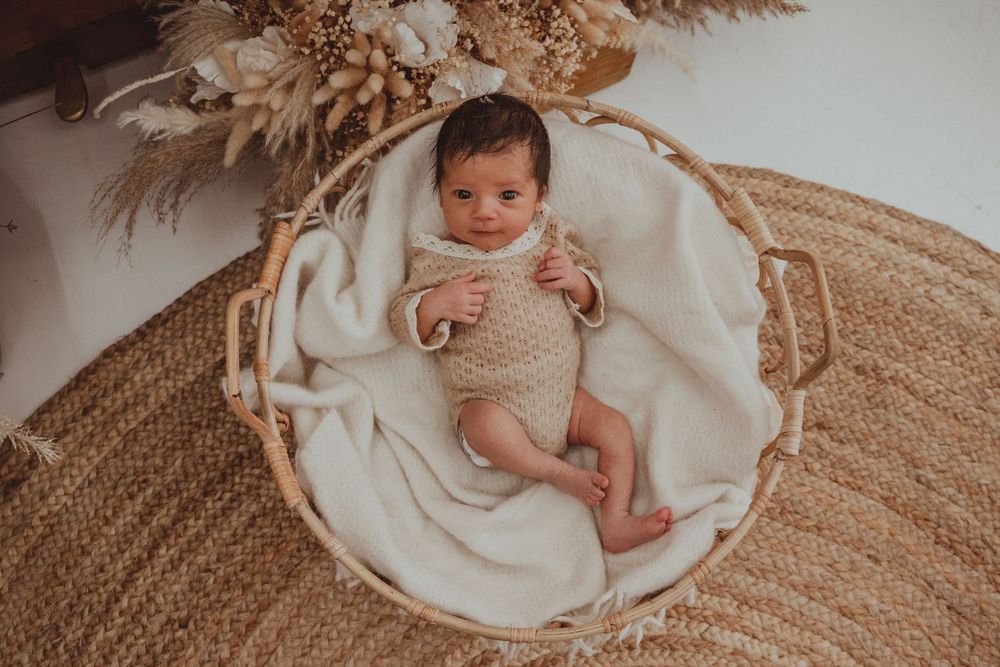 A small infant lies on a soft white blanket in a round woven basket on a beige jute rug.