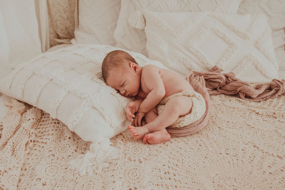 Sleeping newborn baby curled up on white textured bedding in warm natural light.