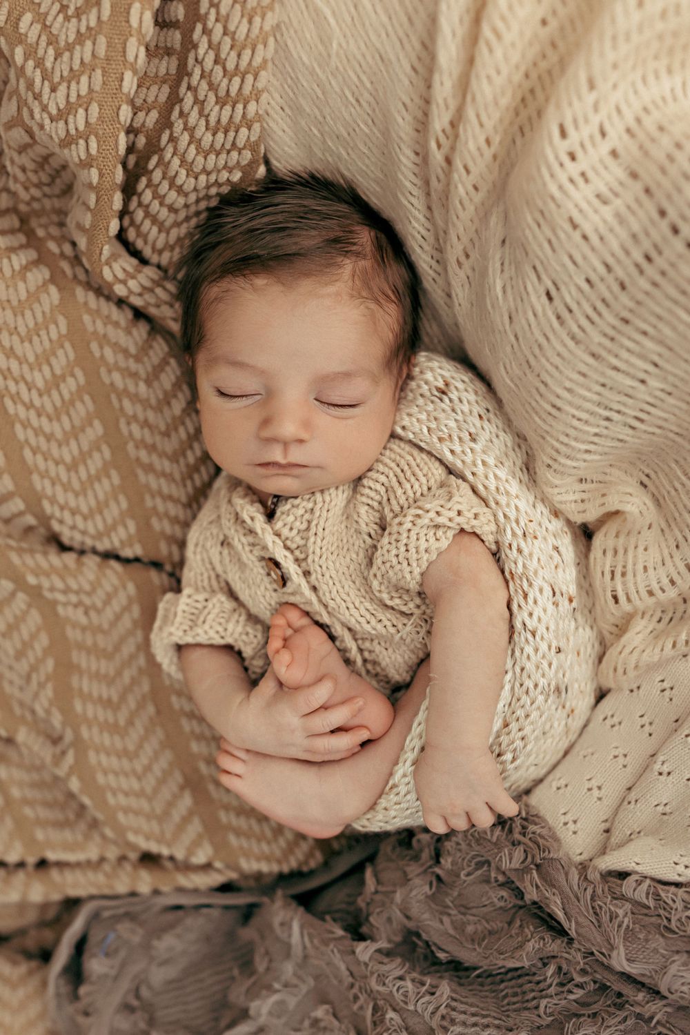 Sleeping newborn wrapped in beige knit blankets on textured neutral backdrop.