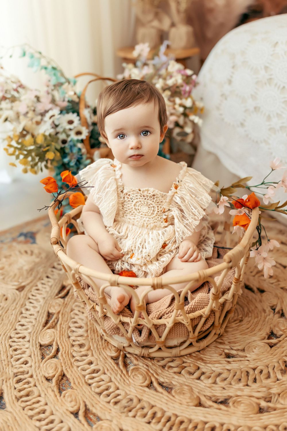 Baby wearing lace dress sits in woven basket surrounded by dried flowers on textured rug.