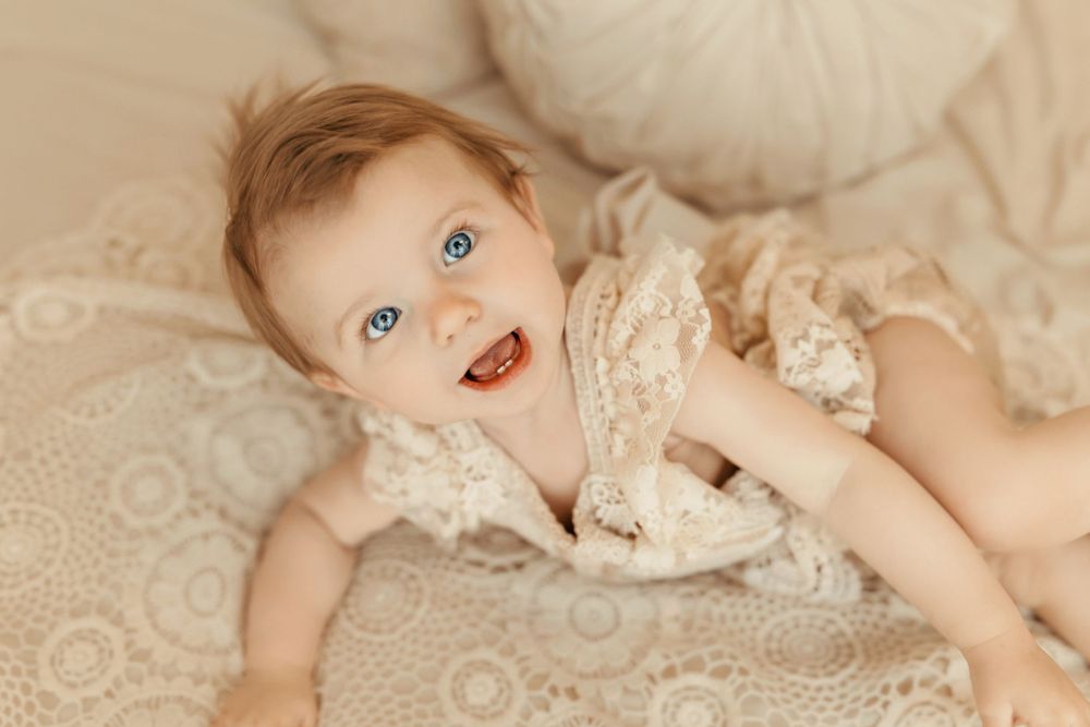 Baby wearing lace clothing lies on cream colored bedding while smiling at camera with bright blue eyes.