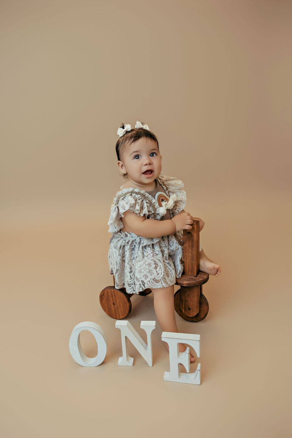 An infant wearing a floral dress sits on a wooden tricycle next to white decorative letters spelling 'ONE' on beige backdrop.