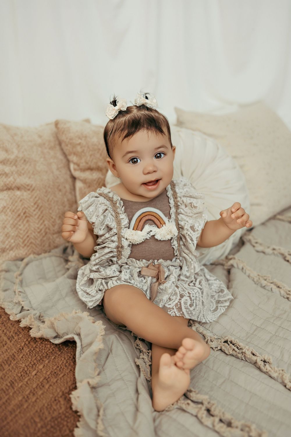 Baby wearing a ruffled beige romper with rainbow design lies on textured beige bedding with a sweet expression.
