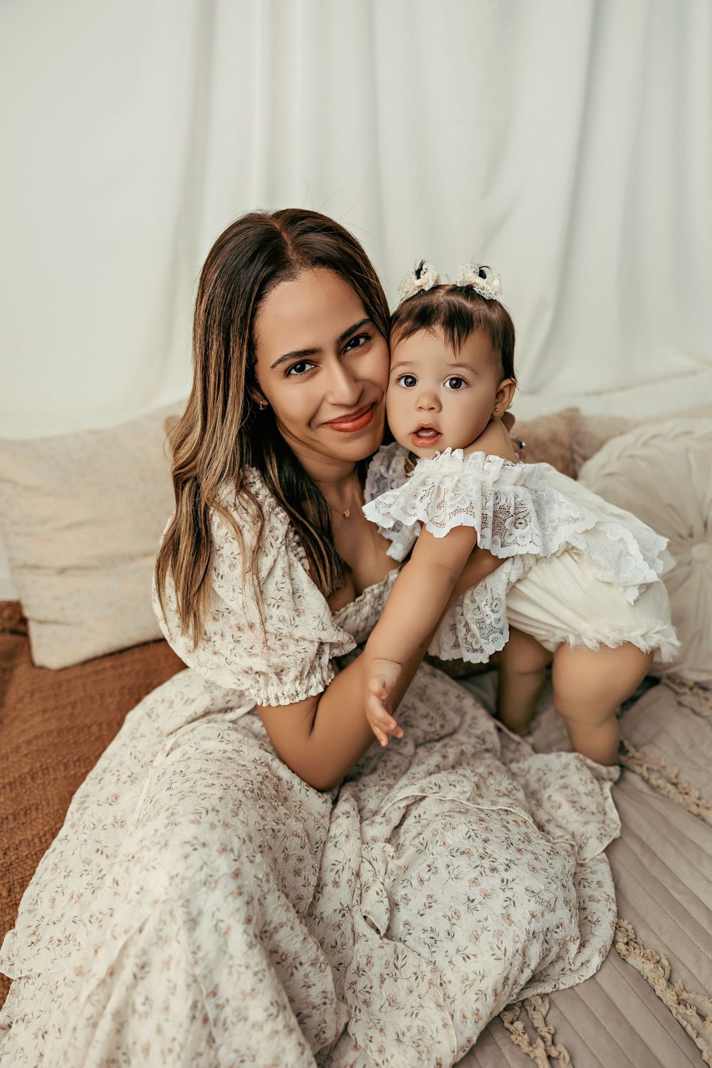 Mother and baby wearing matching white lace dresses pose together on a couch in warm, neutral tones.