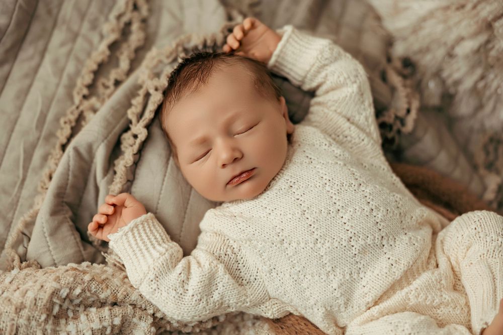 Sleeping newborn baby wearing cozy white knit sweater rests peacefully on textured beige blanket.
