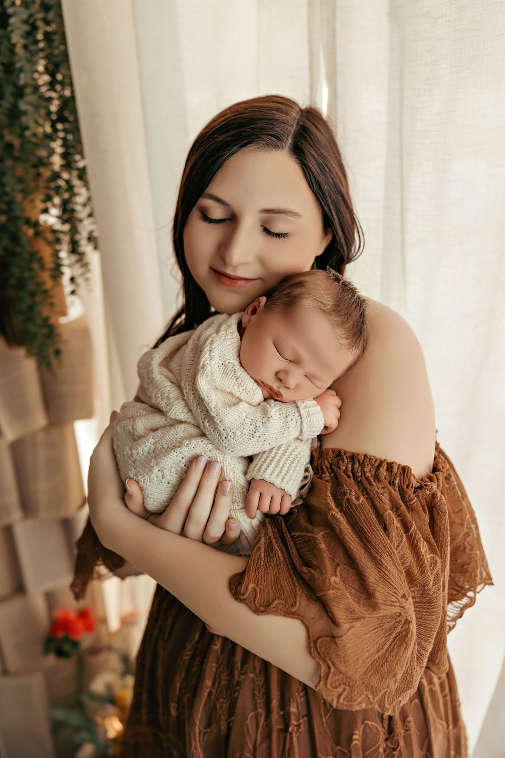 A tender moment captured as a mother in a brown dress holds her sleeping newborn wearing a white knit outfit.