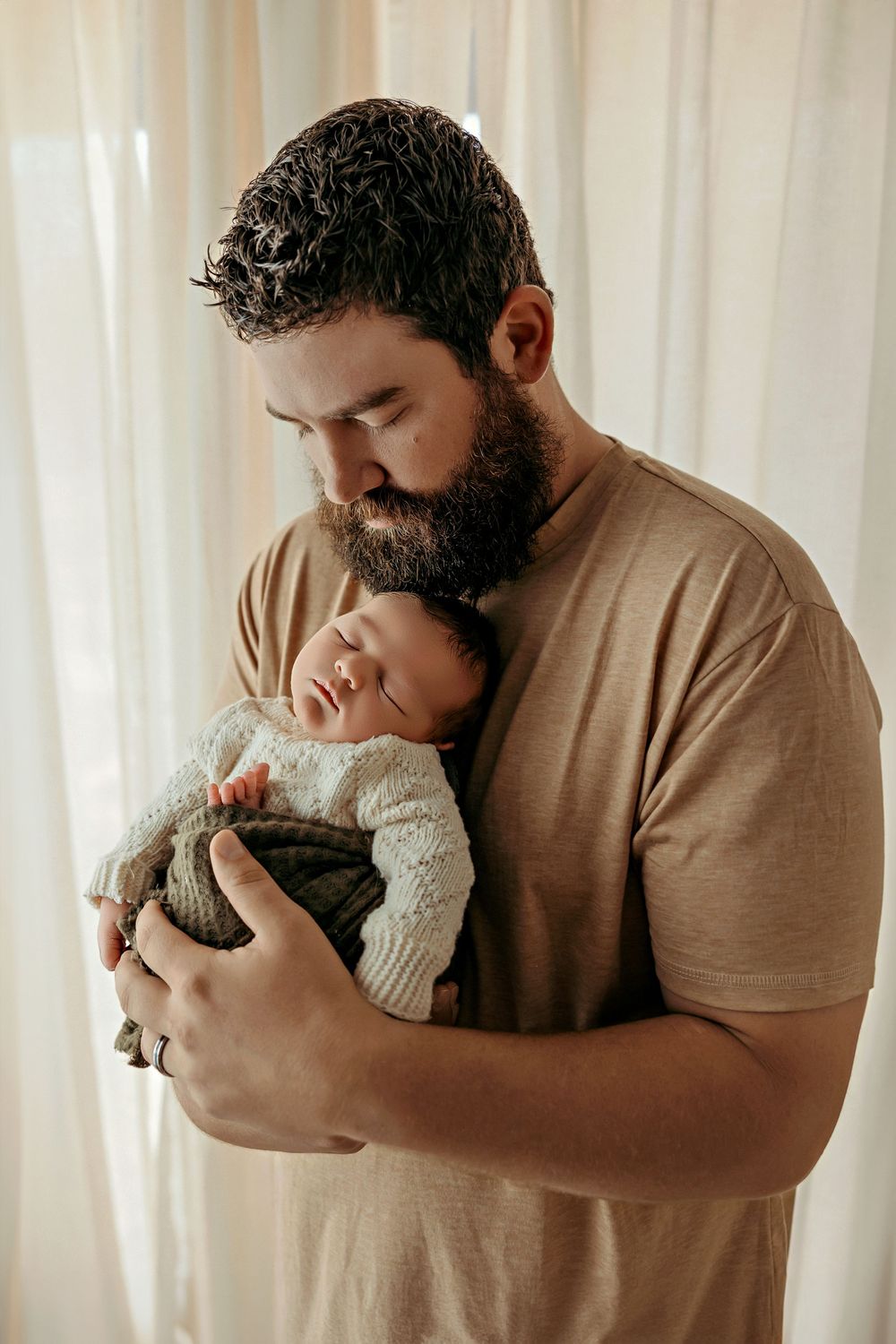 A tender moment captured as a bearded man in a tan t-shirt cradles and kisses a sleeping newborn baby against soft curtains.