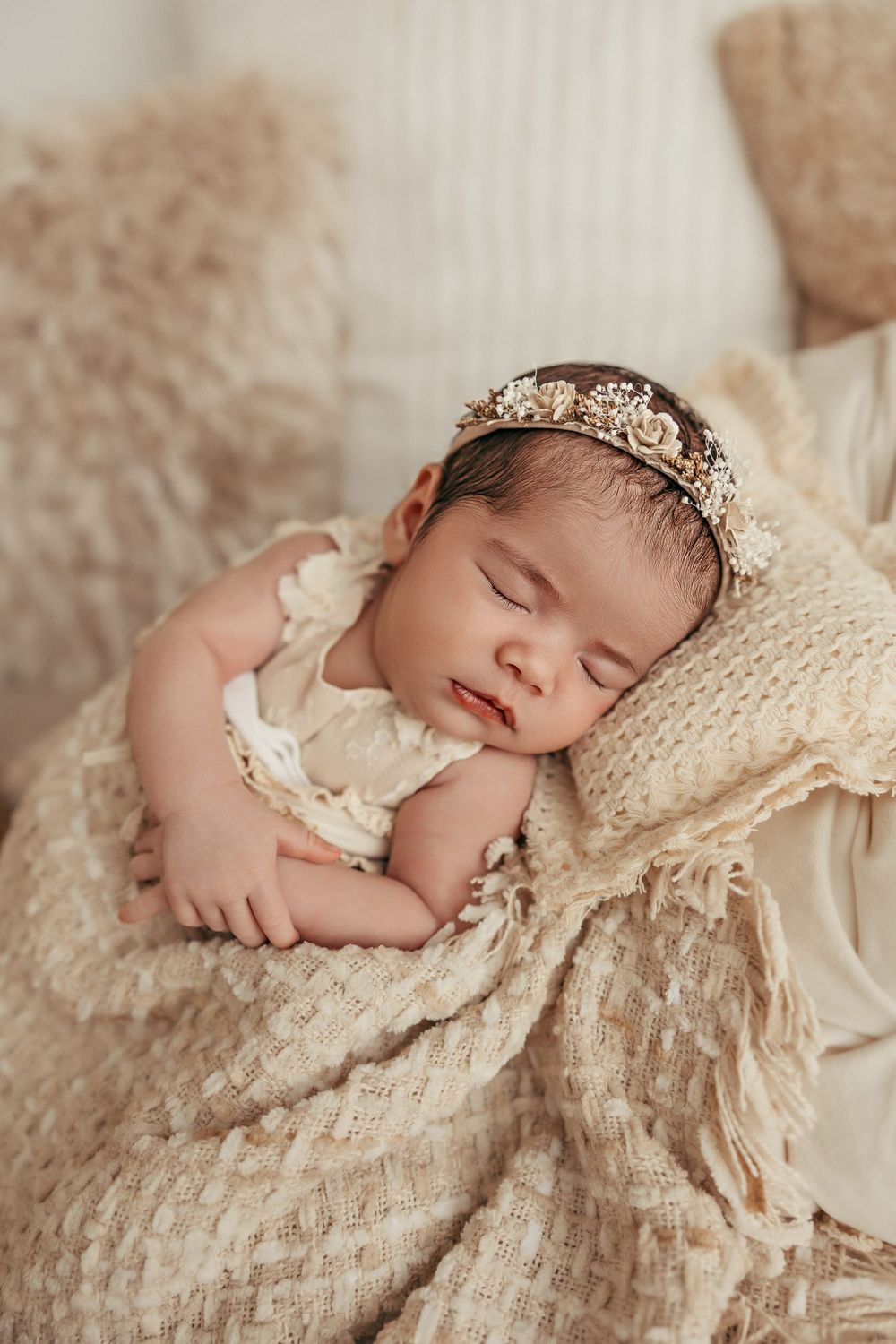 A sleeping newborn wearing a delicate floral headband rests peacefully on cream-colored textured fabric.
