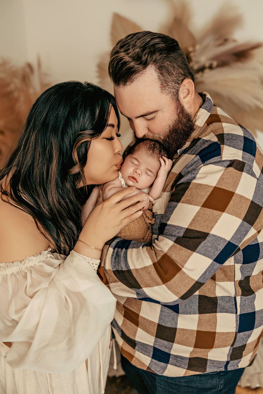 A couple shares an intimate moment with their newborn baby in a warm-toned portrait against a neutral background.