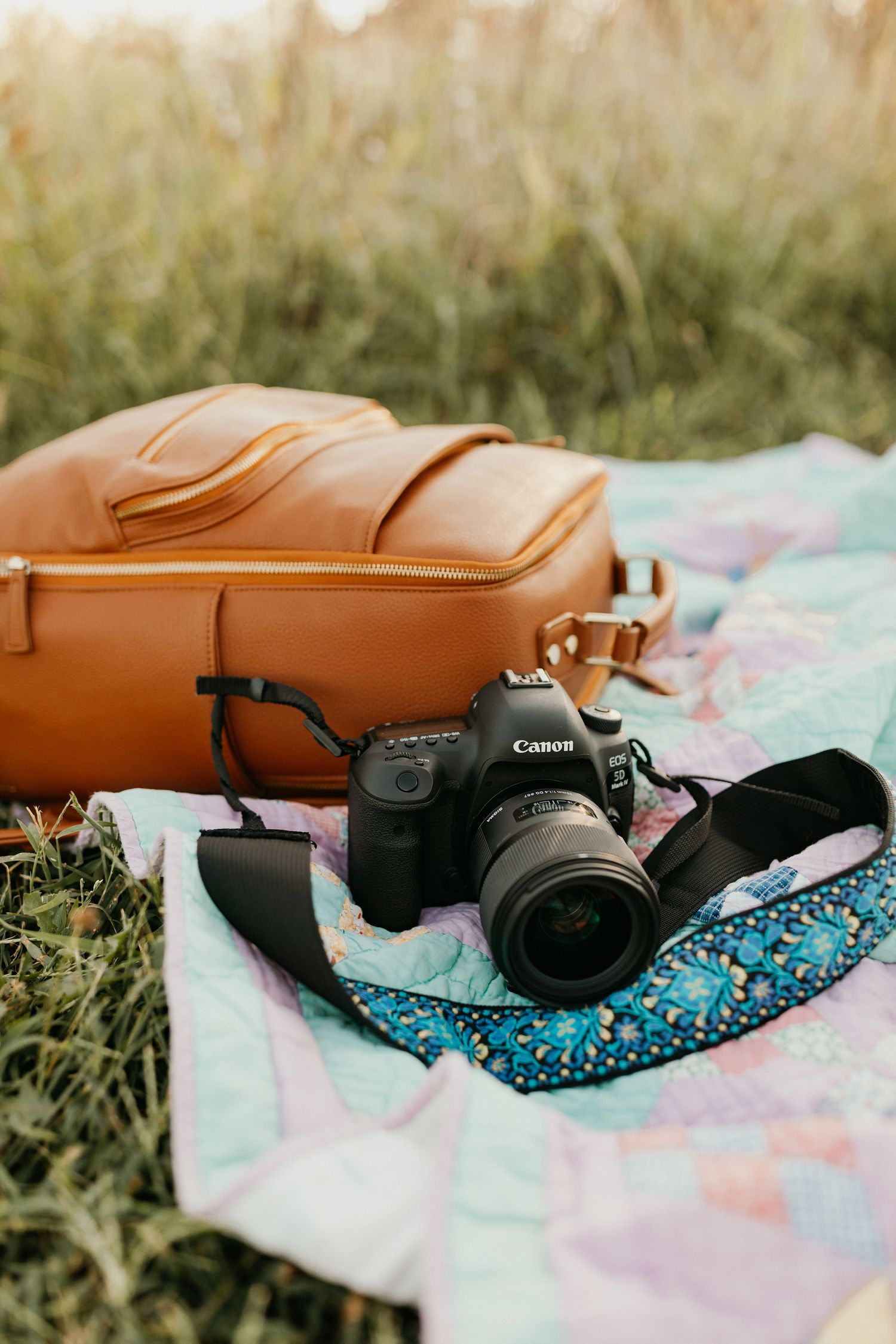 A Canon camera with lens sits on a pastel blanket next to a brown leather bag in tall grass during golden hour.