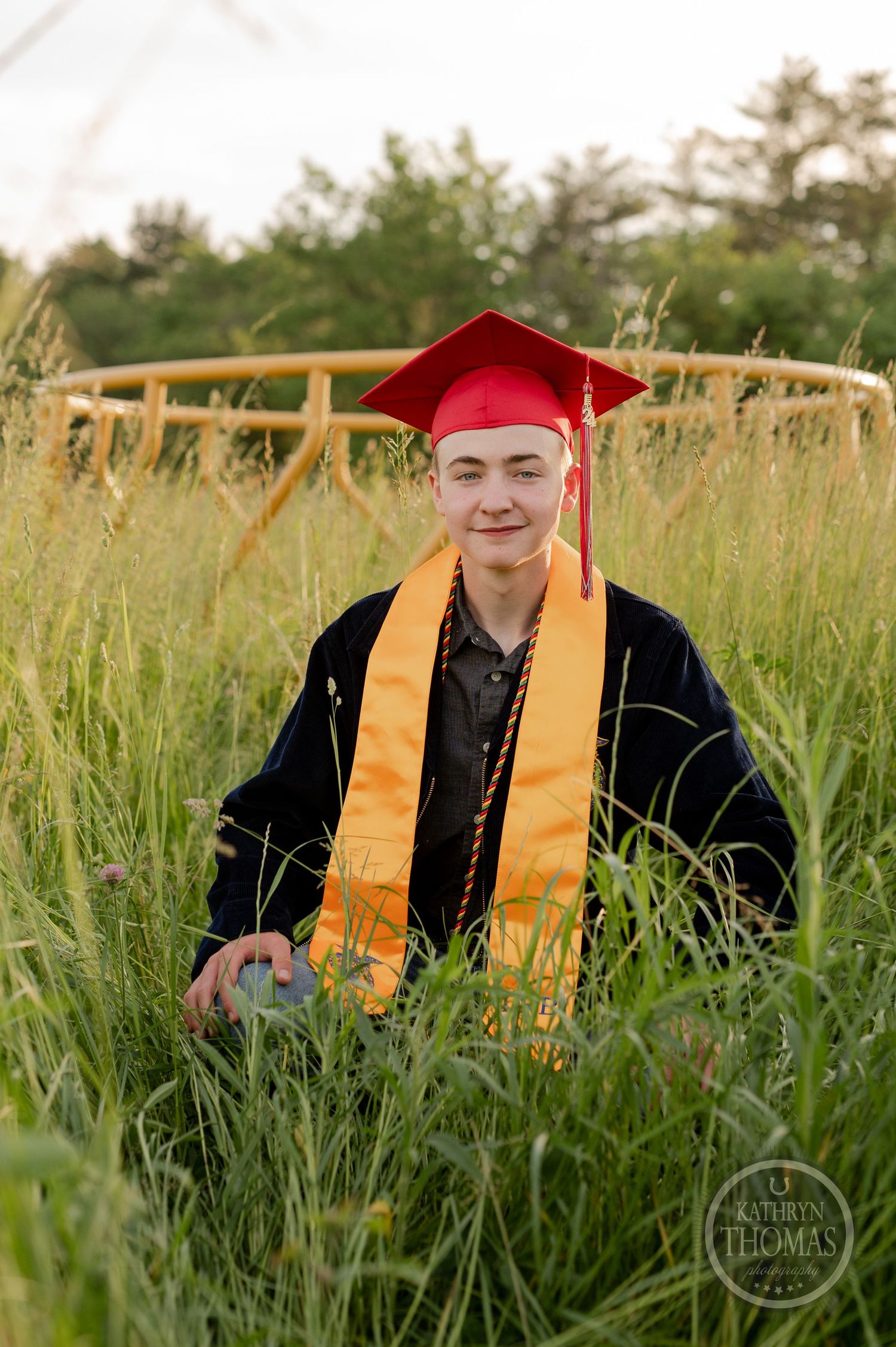 Graduate in red cap and gold stole kneels in tall grass during outdoor photo session.