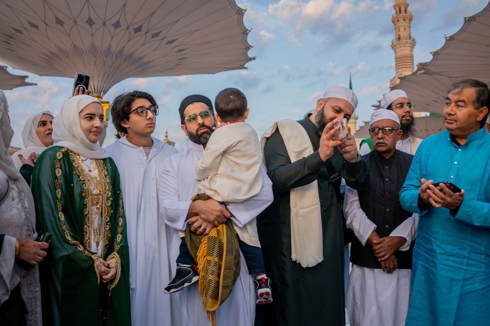 People gather for prayer at a mosque courtyard under a blue sky with architectural details in the background.