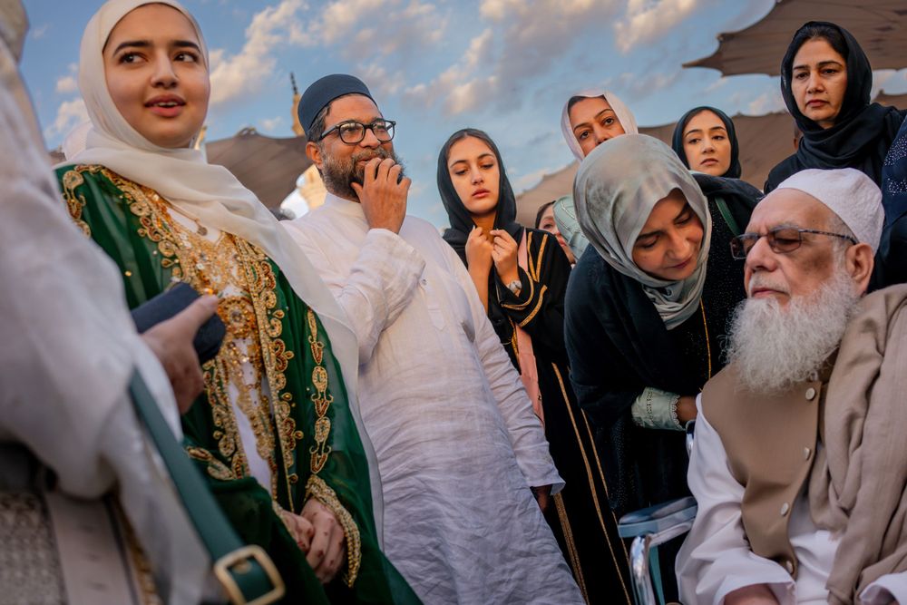A diverse group wearing traditional Muslim attire gathered outdoors against a cloudy sky background.